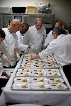 several chefs are preparing food on trays at a table in a kitchen with white linens