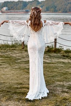 a woman in a white wedding dress with her arms spread out to the side, standing on grass near water