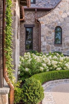 a stone house with white flowers in the front yard and green bushes on either side