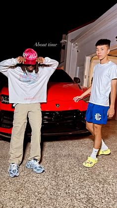 two young men standing next to a red sports car with their hands on their head