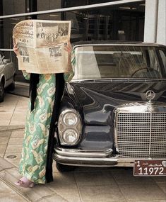 a woman standing next to a black car and holding a newspaper in front of her face