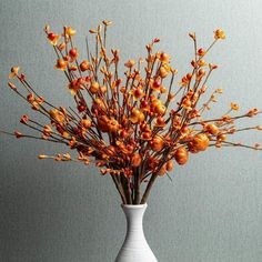 a white vase filled with orange berries on top of a wooden table next to a gray wall