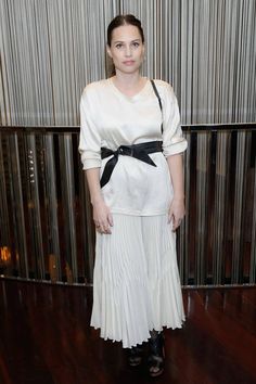 a woman standing in front of a wall wearing a white shirt and pleated skirt