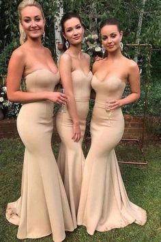 three beautiful women in long dresses posing for the camera on grass with trees and bushes behind them