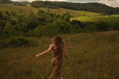 a woman standing in a field with her arms out to the side, looking at trees and rolling hills