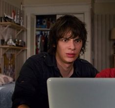 two young men sitting in front of a laptop computer looking at the camera while talking