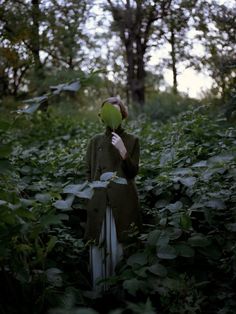 a woman standing in the middle of a forest holding a green object up to her face