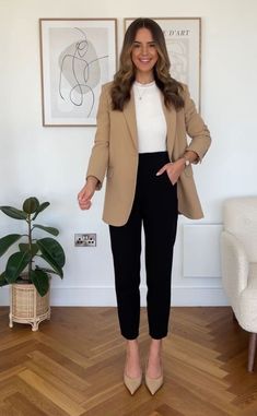 a woman standing in front of a wall wearing black pants and a tan blazer