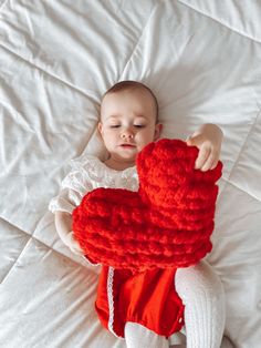 a baby laying on top of a bed holding a red knitted heart shaped object