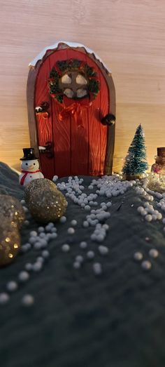 there is a small snowman sitting in front of a red door and some rocks