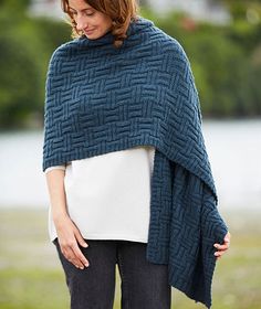 a woman wearing a blue knitted ponchy standing in front of a body of water