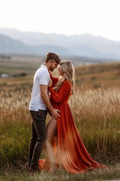 a man and woman standing next to each other in tall grass with mountains in the background