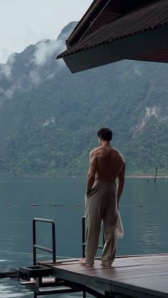 a shirtless man standing on a dock looking out at the water and mountains in the distance