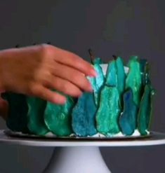 a person is decorating a cake with green frosting and blue icing on a white pedestal