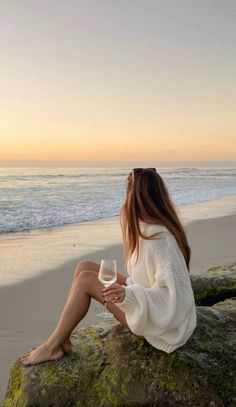 a woman sitting on top of a rock next to the ocean holding a glass of wine