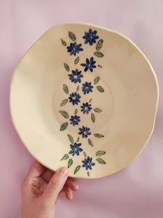 a hand holding a white plate with blue flowers and green leaves on the rim, against a pink background