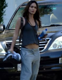 a woman standing in front of a black car holding a water bottle and cell phone