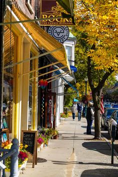 people walking down the sidewalk in front of shops on a sunny day with autumn leaves