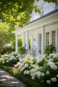 a white house with lots of flowers in the front yard and bushes around the porch