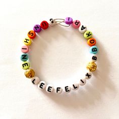 "Stray Kids Individual Member Multi-coloured Alphabet Bead Bangle - Felix. It says \"Lee Felix\" in the centre in white beads, flanked by 2 round golden filigree spacer beads and then 1 of Felix's nicknames and his SKZOO: \"Sunshine\" on the left and \"Bbokari\" on the right in multicoloured beads. The start of each flanking word is also highlighted by a white letter. *Please note that design of spacer beads will vary between bracelets, so no two bracelets will be exactly the same. The bangle it Adjustable White Name Bracelet With Gold Beads, Lee Know Bracelet, Gold Beaded Bracelets With Letter Beads For Festival, Gold Letter Beads Bracelets For Festival, Gold Festival Bracelets With Letter Beads, Gold Bracelets With Letter Beads For Festivals, White Beaded Round Name Bracelet, White Beaded Bracelets With Gold Beads For Party, White Beaded Bracelets With Gold Beads For Festivals