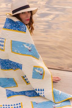 a woman wearing a hat and holding a blue and white blanket over her head while sitting on the edge of a body of water