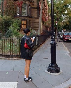 a woman is standing on the sidewalk looking at her cell phone while wearing a leather jacket