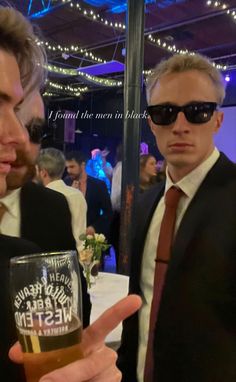 two men in suits and ties holding up a beer glass at an event with lights on the ceiling