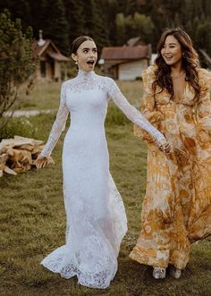 two women in long dresses standing next to each other on the grass with trees in the background