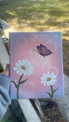 a person holding up a painting with daisies and a butterfly on it in their hand