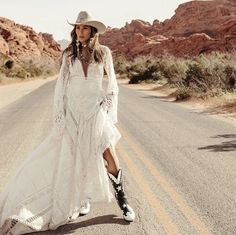 a woman in a long white dress and cowboy hat is walking down the middle of an empty road