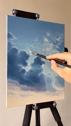 a person holding a paintbrush in front of a painting on a easel with clouds painted on it