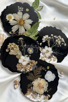 three black plates with white flowers and green leaves on them sitting on a white cloth