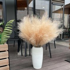 a white vase filled with lots of dry grass on top of a wooden floor next to tables