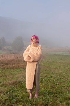 a woman with pink hair standing in a foggy field wearing a sweater and pants