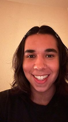 a young man with long hair smiles at the camera while wearing a black t - shirt