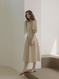 a woman in a beige dress is standing against a wall