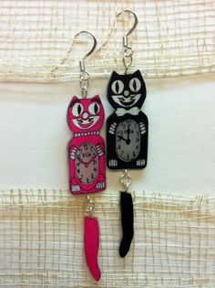 two cats are hanging from earrings on a white surface with a black cat and pink cat