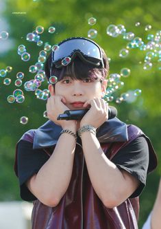 a boy wearing sunglasses blowing bubbles in the air with his hand on his chin and looking at camera