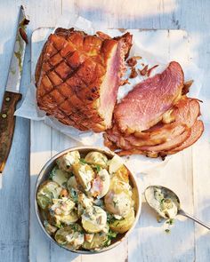 ham and potato salad on a wooden table with a knife next to the dish, along with a large piece of meat