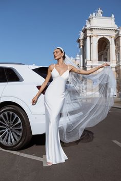 a woman in a white wedding dress standing next to a car with a veil over her head