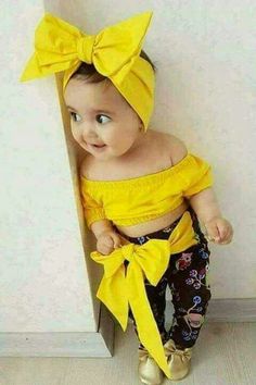 a baby with a yellow bow on her head is standing in front of a wall