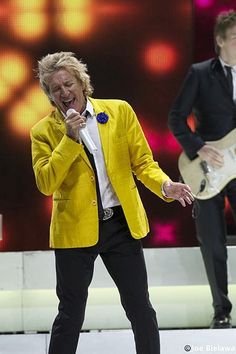 two men in yellow jackets and black pants singing on stage with one holding a guitar