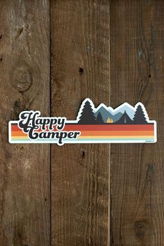 a sticker that says happy camper with mountains and trees in the background on a wooden surface