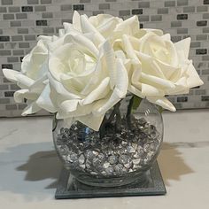 two white roses are in a clear vase on a counter top with rocks and gravel