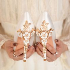 a woman holding two pairs of white shoes with gold leaves and flowers on the heels