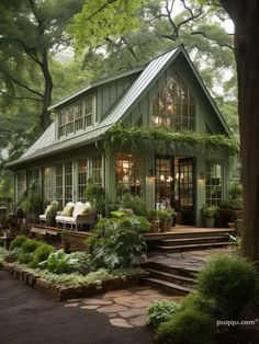 a small green house with lots of windows and plants on the front porch, surrounded by trees