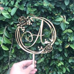 a hand holding a cake topper in front of some green leaves and bushes with the letter a on it