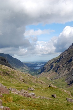 two horses grazing on the side of a mountain with water in the distance and clouds overhead