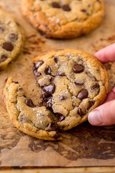 a person holding a chocolate chip cookie in front of other cookies on a baking sheet