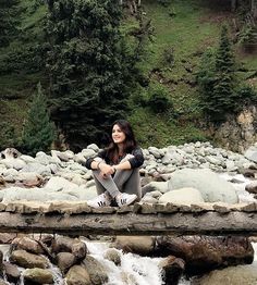 a woman is sitting on a rock bridge over a stream with rocks and trees in the background